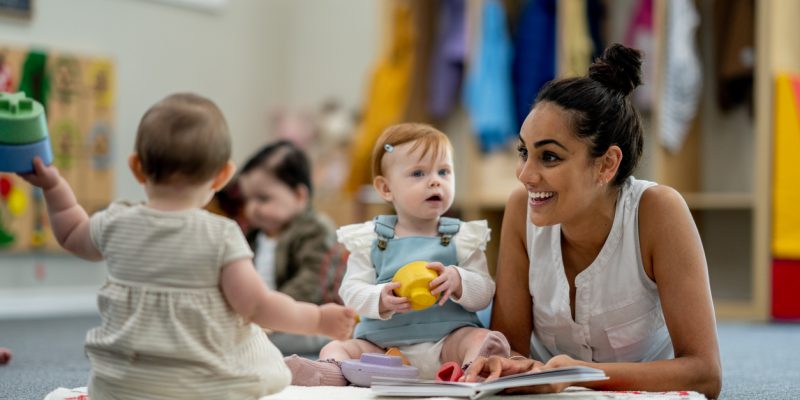 A small group of babies sit on the floor playing with various toys while their teacher supervises and engages them.  They are each dressed casually  and appear content.