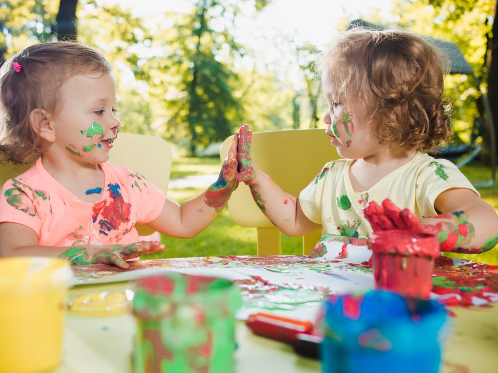 Two-year old girls painting with poster paintings and sitting at a table together against green lawn